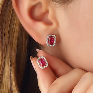 May include: A pair of stud earrings featuring a rectangular, vibrant red center stone surrounded by a halo of small, clear, sparkling white stones set in a metallic frame. One earring is worn on the earlobe, while the other is held by fingers.