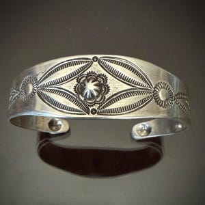 May include: A silver cuff bracelet featuring a central embossed flower motif surrounded by elongated leaf-shaped patterns and smaller circular floral designs. The metal surface has a polished finish with intricate stamped detailing throughout.