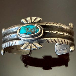 May include: A silver cuff bracelet featuring a central oval turquoise stone with dark matrix patterns. The metal band has a weathered finish with etched diagonal line details and fan-shaped accents above and below the stone, with rounded ends.