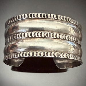 Pode incluir: Uma pulseira larga de prata com um design curvo. A pulseira tem três faixas principais, cada uma com uma superfície lisa e polida. As bordas superior e inferior apresentam um padrão repetitivo de pequenas reentrâncias curvas. A pulseira é aberta na parte inferior.