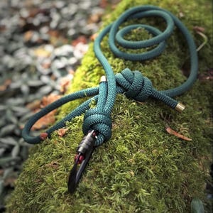 May include: A dark teal rope with a black clasp and gold-colored accents is knotted and coiled on a mossy surface. The rope is thick and appears durable, suggesting a climbing or outdoor activity theme.