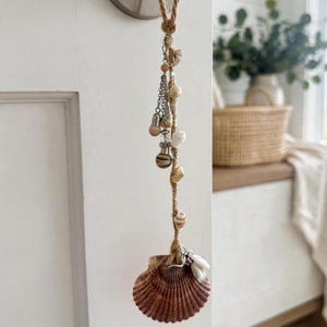 May include: A hanging decorative ornament made of natural jute twine, featuring a variety of small seashells, silver-toned metal charms, and beads. The ornament terminates in a large, dark brown, fan-shaped scallop shell at the bottom end.
