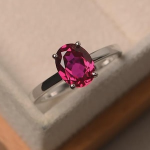 Puede incluir: Un anillo de tono plateado con una gema de color rosa intenso de corte ovalado, sujeta por un engaste de cuatro garras. La banda es lisa y pulida, mostrando un acabado metálico limpio que complementa la vibrante piedra central facetada.
