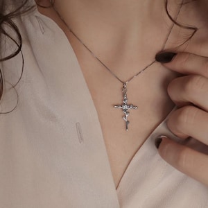 Könnte beinhalten: Eine silberfarbene Halskette mit einem Kreuzanhänger, der ein zentrales Rosenmotiv aufweist und an einer feinen Kette hängt. Der Anhänger zeigt filigrane florale Details entlang der vertikalen und horizontalen Kreuzbalken aus poliertem Metall.