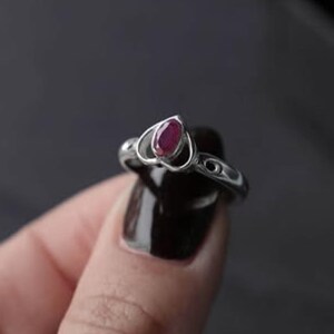 Könnte beinhalten: Ein silberner Ring mit einem polierten, ovalen, tiefroten Edelstein, der in einen herzförmigen Metallrahmen eingefasst ist. Die Ringschiene hat eine glatte, metallische Oberfläche mit dezenten dekorativen Aussparungen an den Seiten.