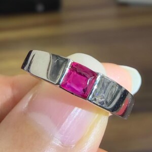 Puede incluir: Un anillo de metal plateado con una piedra preciosa de color rosa vibrante de corte cuadrado en el centro, flanqueada por dos piedras transparentes de corte rectangular engastadas en una banda lisa y pulida de diseño moderno.