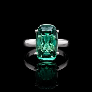 May include: A silver-toned ring featuring a large, rectangular, cushion-cut green gemstone set with four prongs. The stone displays a faceted surface with deep emerald hues and light reflections, mounted on a smooth, polished metal band.