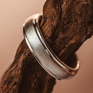 Puede incluir: Un anillo de metal plateado que presenta una banda central ancha y texturizada con un acabado mate y granulado, flanqueada por dos bordes lisos y pulidos que reflejan la luz, rodeando un trozo de madera marrón oscura, rugosa y desgastada.
