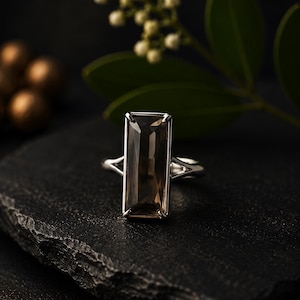 Peut inclure: Une bague en argent ornée d'une grande pierre précieuse rectangulaire en quartz fumé à facettes, fixée par quatre griffes. L'anneau présente un design divisé qui se connecte à la base du sertissage, avec une finition métallique polie.