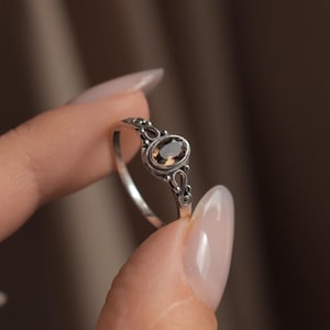 Op de afbeelding: Een zilveren ring met een ovaalvormige, doorschijnende bruine edelsteen in een decoratieve metalen zetting met ingewikkelde kruldetails langs de band. Het metaal heeft een donkere, geoxideerde afwerking die de sierlijke patronen accentueert.