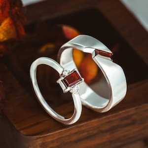 Op de afbeelding: Twee zilveren ringen met dieprode, rechthoekige edelstenen. De dunnere ring heeft een centrale baguette-geslepen rode steen geflankeerd door twee kleine heldere stenen. De bredere band heeft een enkele, verzonken rechthoekige rode steen.