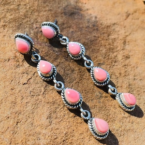 Könnte beinhalten: Ein Paar lange Ohrhänger mit vier tropfenförmigen rosa Steinen in oxidiertem Silber. Jeder Stein ist von einem gedrehten Metallrand umgeben, und die Steine sind durch kleine silberne Verbindungsringe in einer vertikalen Linie angeordnet.