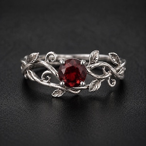 Op de afbeelding: Een zilverkleurige ring met een ronde, dieprode edelsteen in het midden. De band is ontworpen met ingewikkeld, verhoogd metaalwerk dat kronkelende wijnranken en kleine, gedetailleerde blaadjes langs de zijkanten van de steen weergeeft.