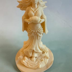 May include: An off-white figurine of a standing figure with long flowing hair, wearing a draped garment with beaded accents, holding a shell in both hands. The figure stands on a circular base featuring sculpted wave-like patterns at the bottom.