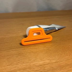 Puede incluir: Un exprimidor de tubos naranja con una abertura rectangular y dos ranuras, apoyado sobre una superficie de madera. Un tubo de producto parcialmente usado está insertado en el exprimidor. El exprimidor está diseñado para ayudar a extraer el contenido del tubo.