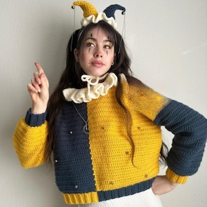 May include: A handmade crochet jester costume featuring a color-blocked sweater in navy blue and yellow, adorned with silver studs. The outfit includes a matching jester hat and a cream-colored ruffled collar. The overall aesthetic is whimsical and theatrical.