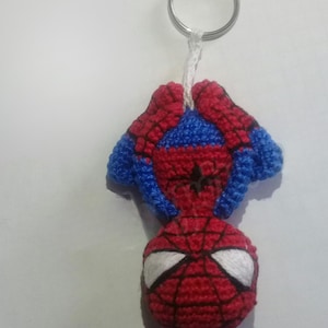 Op de afbeelding: Een handgemaakte gehaakte Spider-Man sleutelhanger. De sleutelhanger heeft een rood en blauw Spider-Man ontwerp met witte ogen en een zwart webpatroon. Het is bevestigd aan een zilveren metalen sleutelring met een witte draad.