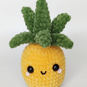 Könnte beinhalten: Ein gehäkeltes Ananas-Spielzeug mit einem lächelnden Gesicht. Die Ananas ist gelb mit einer strukturierten Oberfläche, grünen Blättern und schwarzen Augen. Kleine rosa Akzente befinden sich unter den Augen.
