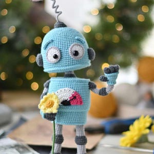Op de afbeelding: Een handgemaakte gehaakte robot in lichtblauw, grijs en bruin. De robot heeft een hartvormige kopbevestiging en houdt een gele bloem vast. Het lichaam van de robot heeft een waaiervormig ontwerp. Een grillig, handgemaakt item.