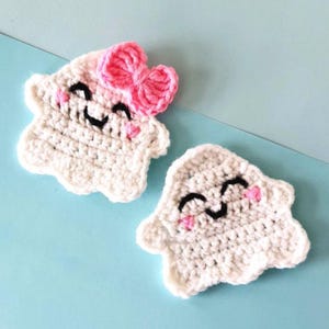 May include: Two handmade crochet ghost appliques. One ghost has a pink bow, black eyes, and pink cheeks. The other ghost has black eyes and pink cheeks. Both are white and have a smiling expression.