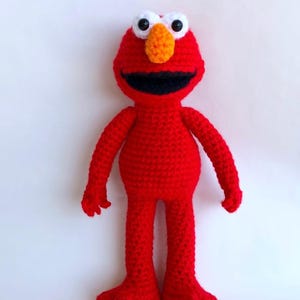 May include: A handmade, red crochet doll with a smiling face. The doll has white eyes with black pupils, an orange nose, and a wide black mouth. The arms and legs are extended, and the doll is standing upright.