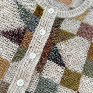 May include: A close-up of a beige, open-knit cardigan with a checkerboard pattern in muted colors. The cardigan has a ribbed collar and button closure with white buttons. The color blocks include shades of brown, green, and blue.