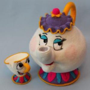 Op de afbeelding: Een gehaakte theepot en kop en schotel set. De theepot is wit met een paars deksel, gele handvat en roze accenten. De kop en schotel is wit met een gele handvat en een rode, blauwe en gele basis. Beide hebben geborduurde gezichtskenmerken.