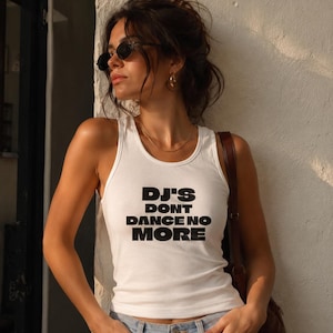 Może przedstawiać: Biały top na ramiączkach z napisem "DJ'S DONT DANCE NO MORE" pogrubioną czcionką. Top zestawiono z jasnoniebieskimi dżinsami. Osoba nosi okulary przeciwsłoneczne i złotą biżuterię.