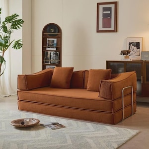 May include: A modern, low-profile sofa in a warm orange-brown hue. The sofa features a rectangular design with thick, cushioned armrests and backrests, complemented by matching throw pillows. The frame has a silver metal accent.