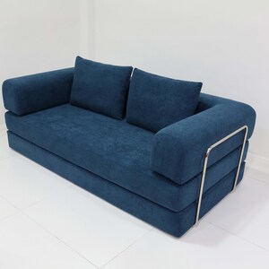 May include: A modern, two-seater sofa in a deep blue fabric. The sofa features two square back cushions and rounded armrests. The frame has a silver metal accent on the side. The sofa sits on a white tiled floor.