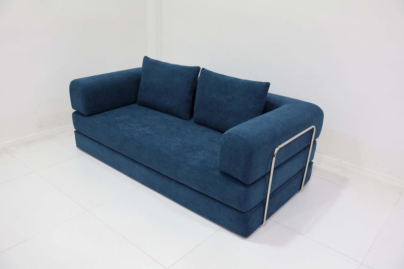 May include: A modern, two-seater sofa in a deep blue fabric. The sofa features two square back cushions and rounded armrests. The frame has a silver metal accent on the side. The sofa sits on a white tiled floor.