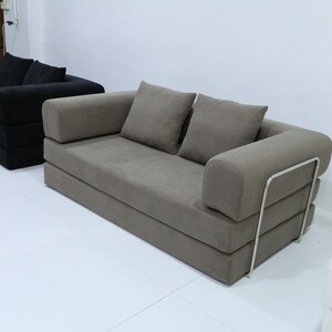 May include: Two modern sofas in a white room. One is a dark gray color, and the other is a light gray color. The light gray sofa has two square pillows and a metal frame on the back.