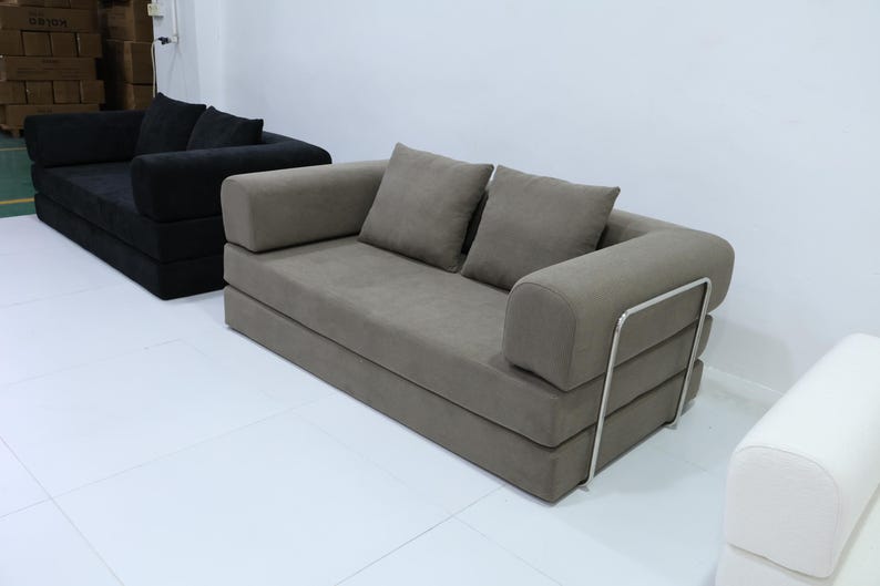 May include: Two modern sofas in a white room. One is a dark gray color, and the other is a light gray color. The light gray sofa has two square pillows and a metal frame on the back.