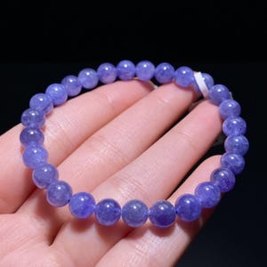May include: A beaded bracelet composed of round, polished, translucent violet-blue gemstone beads strung on an elastic cord. The beads exhibit natural variations in color intensity and internal texture, creating a uniform circular shape.