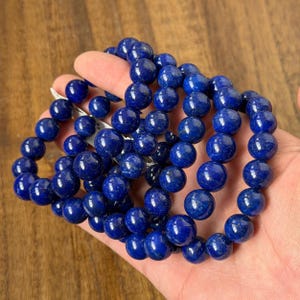 Könnte beinhalten: Mehrere tiefblaue Perlenarmbänder. Jedes Armband besteht aus runden, polierten Perlen. Die Perlen haben eine glatte, reflektierende Oberfläche. Die Armbänder werden in einer Hand gehalten, um ihre Farbe und Textur zu zeigen.
