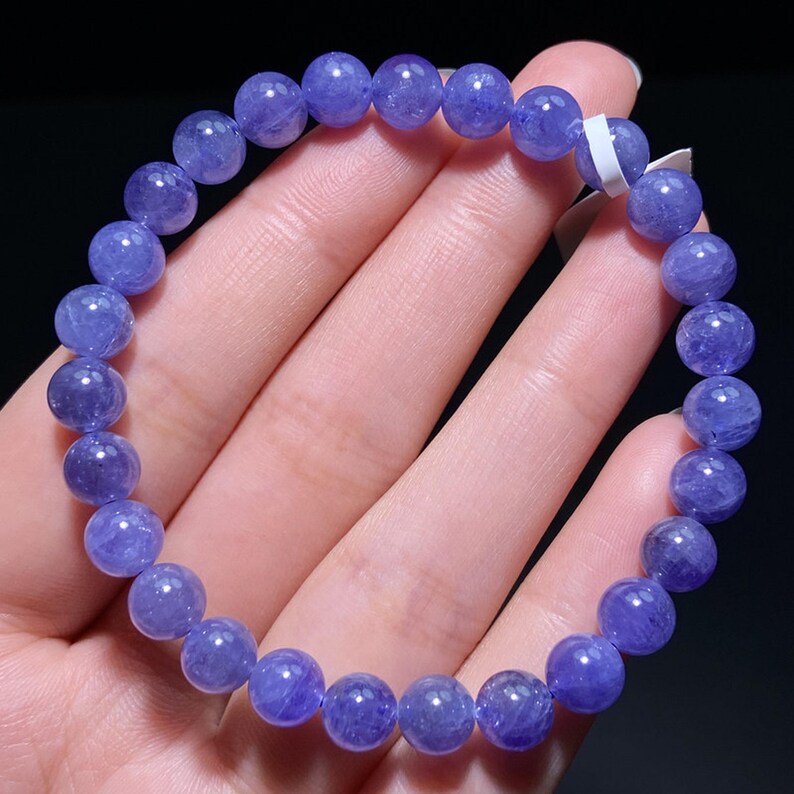 May include: A circular beaded bracelet composed of numerous smooth, round, translucent violet-blue tanzanite beads strung together on an elastic cord. The beads exhibit natural variations in color intensity and internal clarity, with a small white tag.