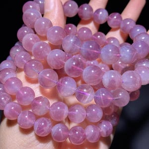 May include: A multi-strand bracelet composed of polished, spherical beads in varying shades of translucent purple and pink. The beads exhibit a smooth, glossy finish with internal inclusions and light-reflecting properties throughout the circular stones.