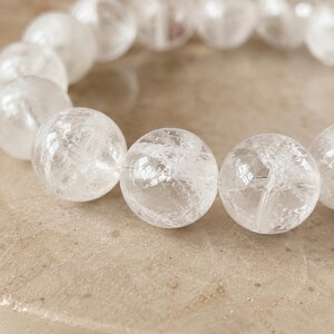 Könnte beinhalten: Ein Armband bestehend aus mehreren kugelförmigen, klaren und durchscheinenden Quarzperlen. Jede Perle hat eine polierte Oberfläche mit sichtbaren inneren Rissen und natürlichen Einschlüssen, die das Licht einfangen und ein helles, reflektierendes Aussehen erzeugen.