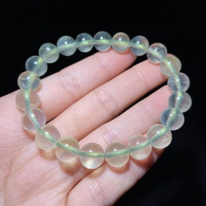 May include: A stretch bracelet composed of round, translucent, pale green beads. The beads have a smooth, polished finish and are strung together on a flexible cord, creating a circular shape that displays subtle internal variations in color.