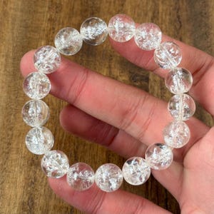 Könnte beinhalten: Ein elastisches Armband aus runden, klaren, rissigen Quarzperlen. Die Perlen haben ein durchscheinendes Aussehen mit inneren Brüchen, die eine reflektierende, eisige Textur auf jedem kugelförmigen Stein erzeugen, aufgereiht auf einer elastischen Schnur.