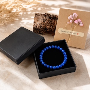 May include: A blue beaded bracelet made of smooth, round, polished stones rests inside a square black gift box. Next to the box is a tan card decorated with a small sprig of dried pink flowers and a sticker that reads Especially for you.