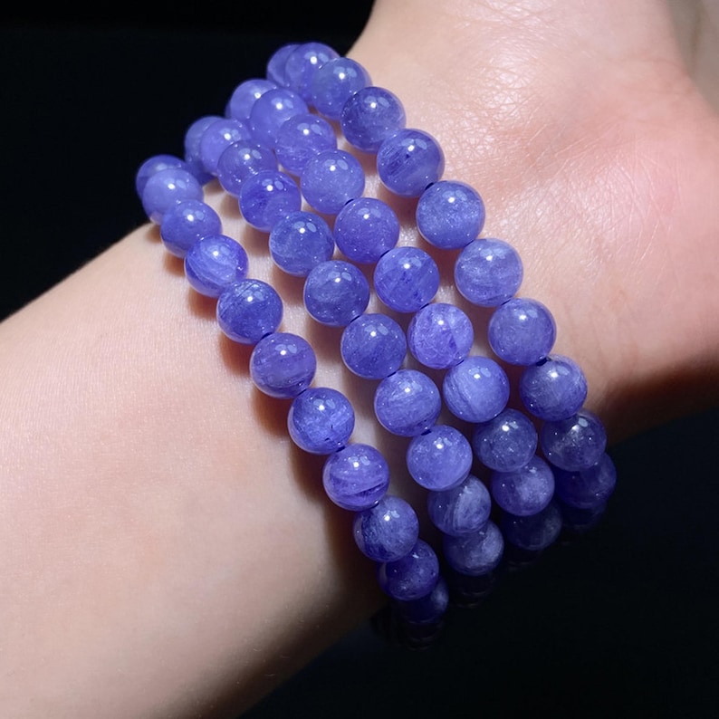 May include: A multi-strand bracelet composed of polished, round, translucent violet-blue beads. The beads feature internal fibrous inclusions and a smooth, glossy finish, arranged in several parallel rows that wrap around the wrist area.