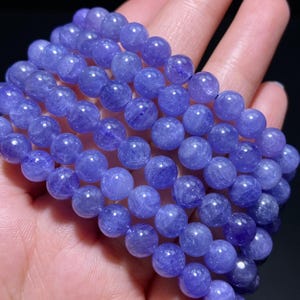 May include: A multi-strand bracelet composed of polished, round, translucent purple beads. The beads exhibit a natural, crystalline texture with varying shades of violet and lavender, arranged in several parallel rows to form a wide, beaded band.