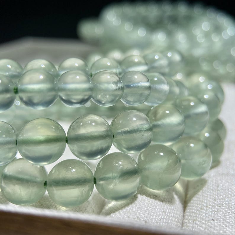 May include: A strand of smooth, round, translucent light green beads strung together. The beads exhibit a soft, pale mint hue with subtle internal variations and a polished, reflective surface, arranged in multiple circular loops.
