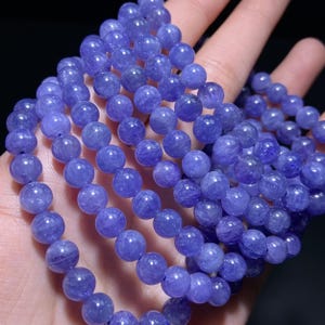 May include: A strand of polished, round, translucent purple beads. The beads exhibit a smooth, glossy surface with subtle internal variations and natural inclusions, reflecting light across their spherical shapes in a uniform, continuous arrangement.