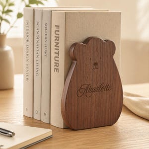 Peut inclure: Un serre-livres en bois brun foncé en forme d'ours, avec un visage gravé simple et le prénom Charlotte en écriture cursive. Le serre-livres soutient trois livres aux tons clairs portant les inscriptions Interior Design Review, Scandinavian Living, Modern Home et Furniture.