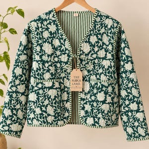 May include: A quilted jacket featuring a dark green floral pattern on a white background, long sleeves, a V-neckline with tie closures, and two front pockets. The interior lining has a green and white vertical stripe pattern. A tag reads THE BLOCK LABEL.