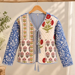 May include: A quilted jacket featuring a V-neckline with blue and white striped trim and tie closures. The front panels display cream fabric with red poppy and floral block prints, while the long sleeves feature a blue and white floral pattern.