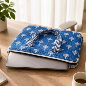 May include: A quilted laptop sleeve in a vibrant blue colour featuring a repeating white floral pattern. The sleeve has two striped fabric handles and a white zipper closure. A silver laptop is partially inserted into the sleeve, revealing its metallic finish.