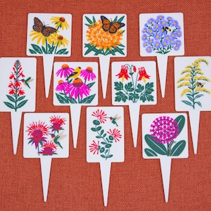 May include: A collection of nine white rectangular plant markers, each featuring a unique colorful illustration of native flowers, butterflies, hummingbirds, bees, and caterpillars, with a pointed stake at the bottom for insertion into soil.